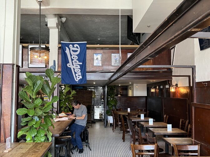 Don Tahara, owner of Far Bar, sits at one of Far Bar's tables and does work. A blue Dodgers banner is draped directly above him.