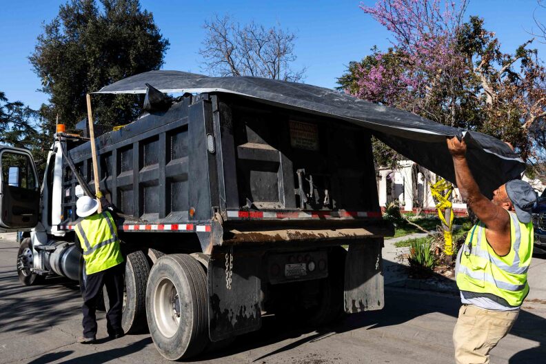 Mean wearing high-visibility vests cover a black dump truck with a black tarp.