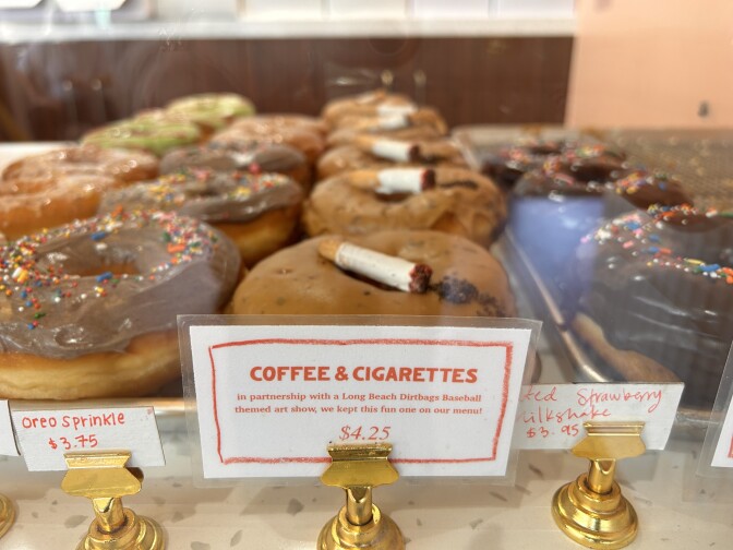 A close-up of a case of doughnuts, with at least three different types lined up next to each other. A small white and orange label reads "coffee & cigarettes ... $4.25" in front of doughnuts with medium-brown icing and fake, edible cigarette placed on top towards the center. 
