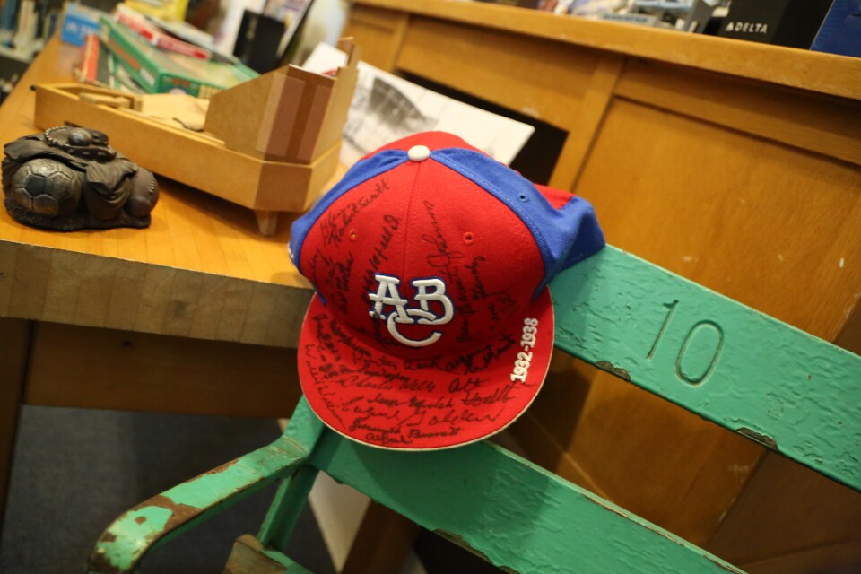 A red and white baseball cap with the letters A B C embroidered in white lettering for its center logo. The cap has multiple autographs in black felt tip marker the entire front.  The cap sits on a light-green stadium chair with the number 10 on it. 