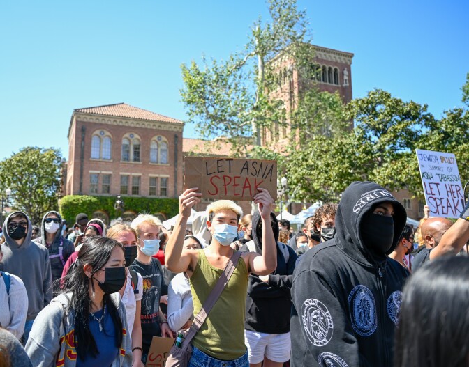 A person wearing an olive green tank top holds up a sign that reads Let Asna Speak. They are surrounded by other people who are mostly masked. 