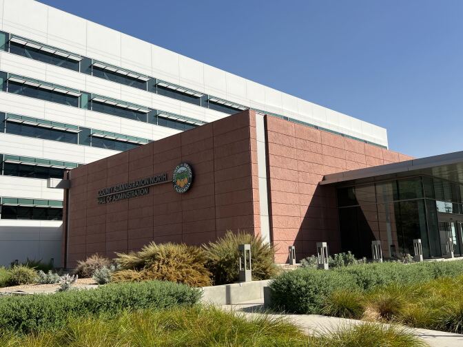 The exterior of a building has a terra cotta-colored stone wall with the words : County of Orange County Administration North 