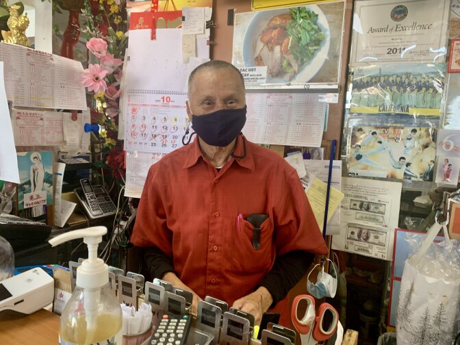 An older Vietnamese American man wearing a red shirt and a black mask poses at the cash register of his family restaurant.