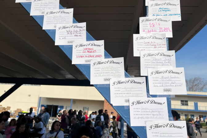 About a dozen certificate-style signs that read I Have A Dream! are taped on a support beam outside a school where dozens of people are gathered on a school playground. Each includes spaces filled in by children that say 'My Name Is,' 'I Am From,' and 'My Dream for the World Is"