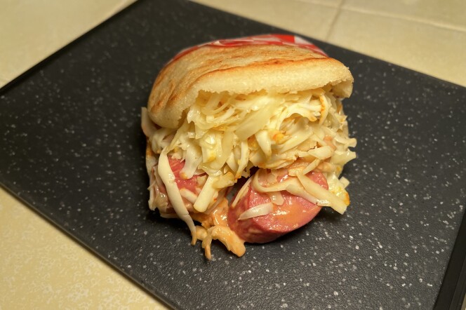 An image of an arepa, made with two white corn flour buns, containing a large pile of white grated cheese dressed with an orange sauce, underneath with sliced pink sausage falling out. The arepa sits on a black-speckled cutting board on a yellow-tiled surface.  