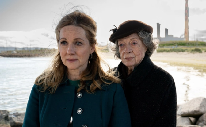 Laura Linney and Maggie Smith stand on a rocky shore on the beaches of Ireland. The sky is grey, and the outline of an industrial plant can be seen in the background. Linney wears a blue coat, the wind gently moving wisps of her hair. She looks in front of her, eyes wide, glints of tears in them. Smith stands right behind her looking in the same direction over her shoulder. She wears a black coat and hat. Her facial expression seems resolute. 