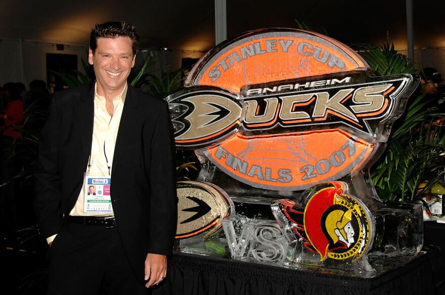 A man with light-tone skin wears a suit jacket and no tie standing next to an Anaheim Ducks Stanley Cup sign