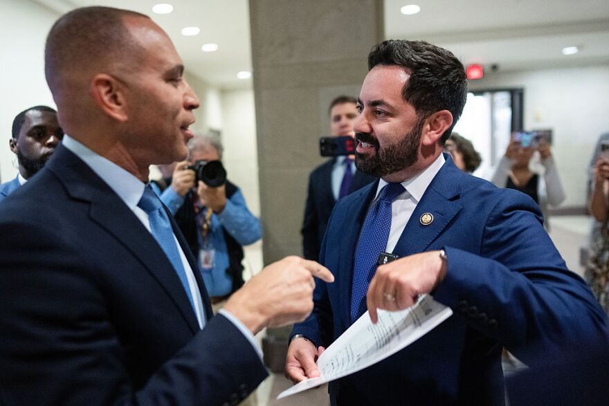 Two man in suits stand close together and both are gesturing to a piece of paper.