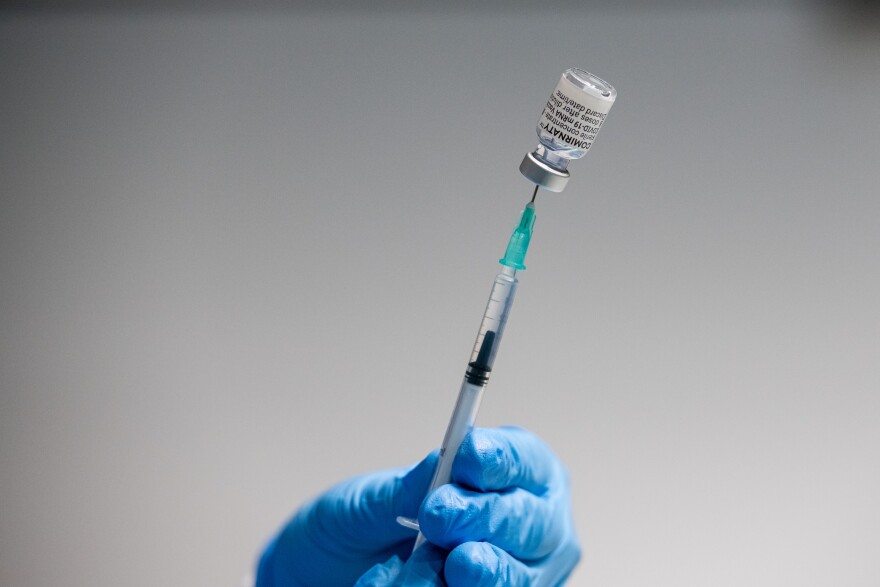 A blue-gloved hand holds a vial of COVID-19 vaccine on the tip of a needle.