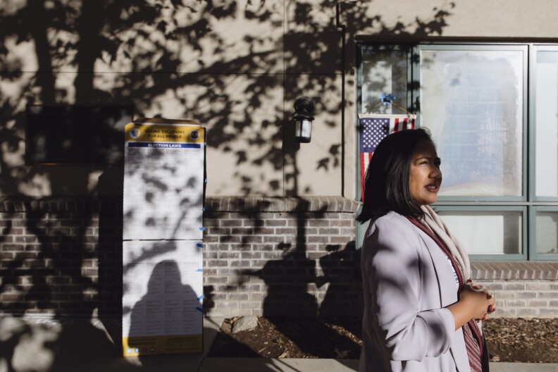 A woman with medium-light skin tone and short dark hair wearing a lavender blazer stands outside an area with dappled light from a tree. Behind her is a notice of election laws near a wall where an American flag hangs.