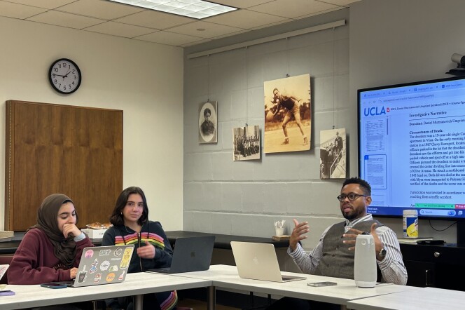Three people sit in a small classroom, with their laptops in front of them. One of the people --- a man with medium skin tone, short dark hair, a long-sleeved shirt, and black-framed glasses-- gestures while explaining something.  Behind him, a document containing the words "Circumstances of Death" is projected on a large screen. 