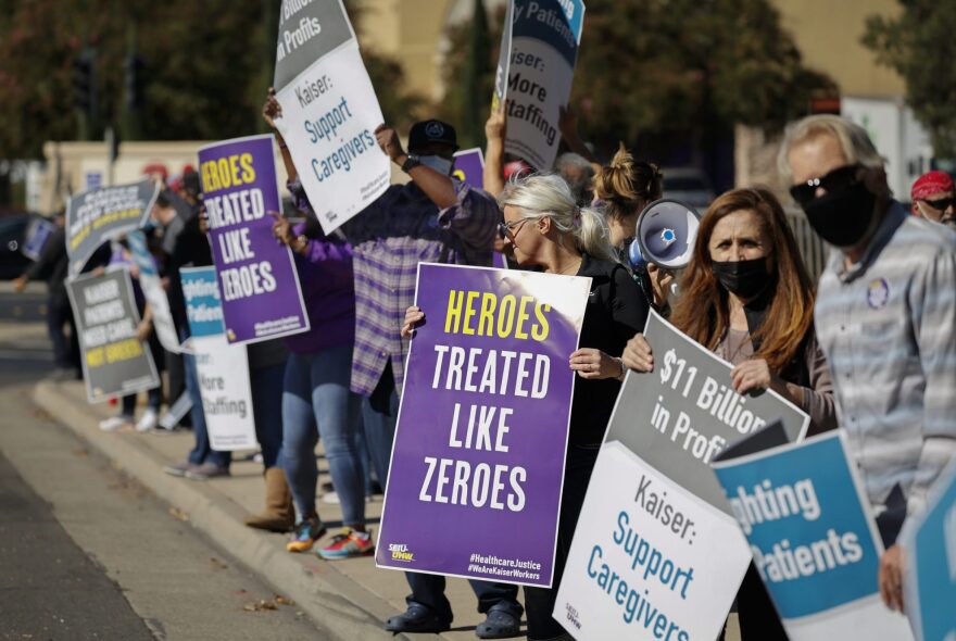 People lining a sidewalk hold ip signs that read "Heroes treated like zeroes" and "$11 Billion in Profits, Kaister Suppor Caregivers." Some are masked and some are not. A bullhorn is visible in the center right.